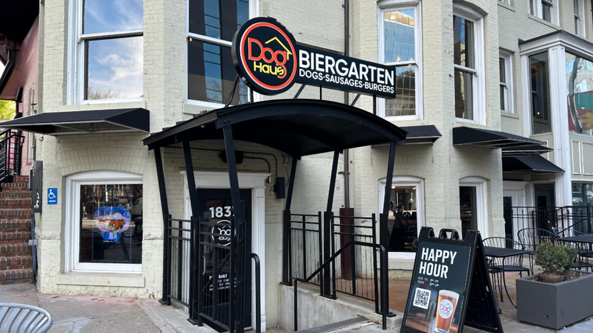 Entrance of Dog Haus Biergarten Dupont with a black awning, branded signage, patio seating, and a sidewalk “Happy Hour” si