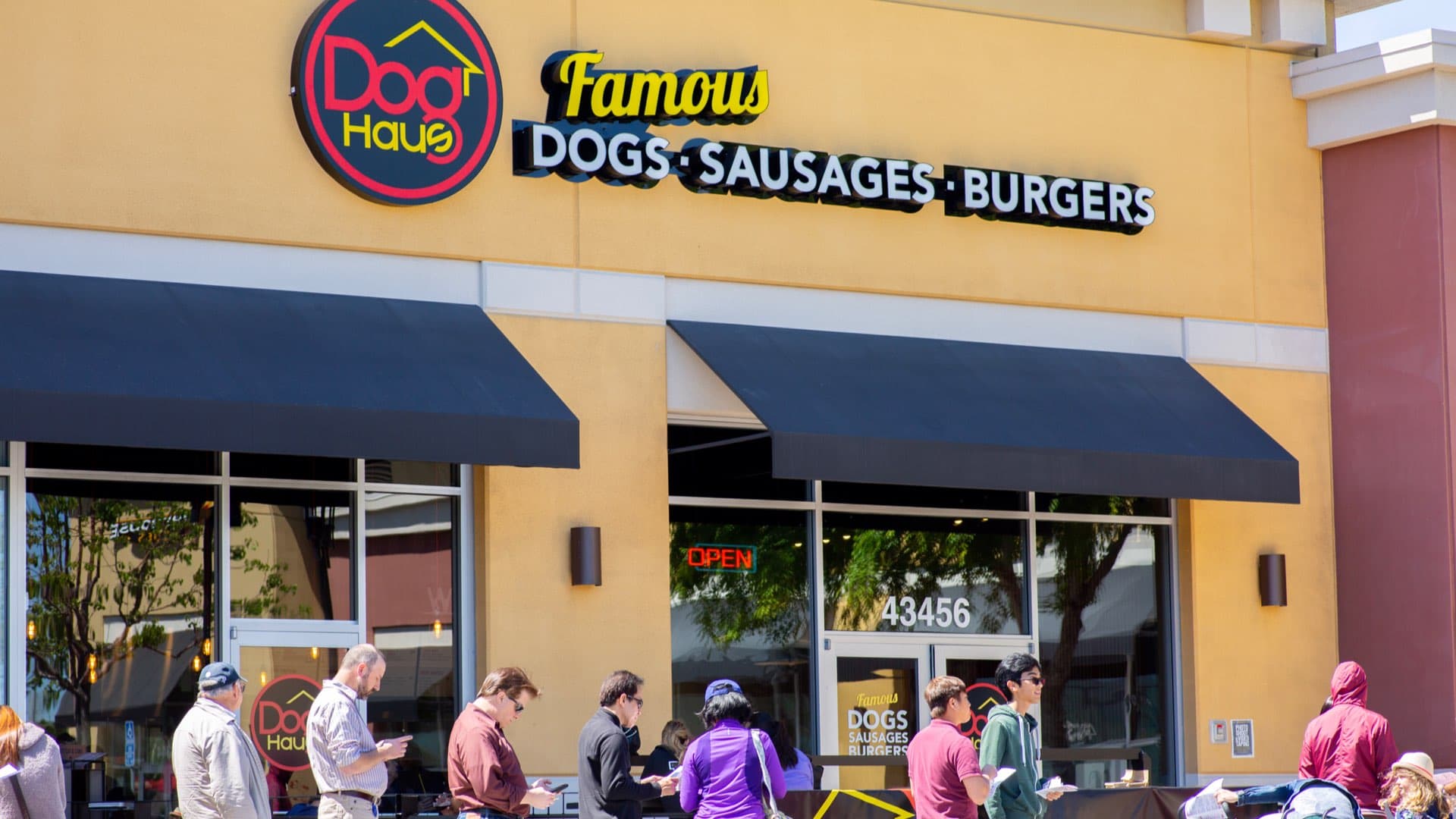 Exterior of Dog Haus Fremont with yellow facade, black awnings, and people in line out front.