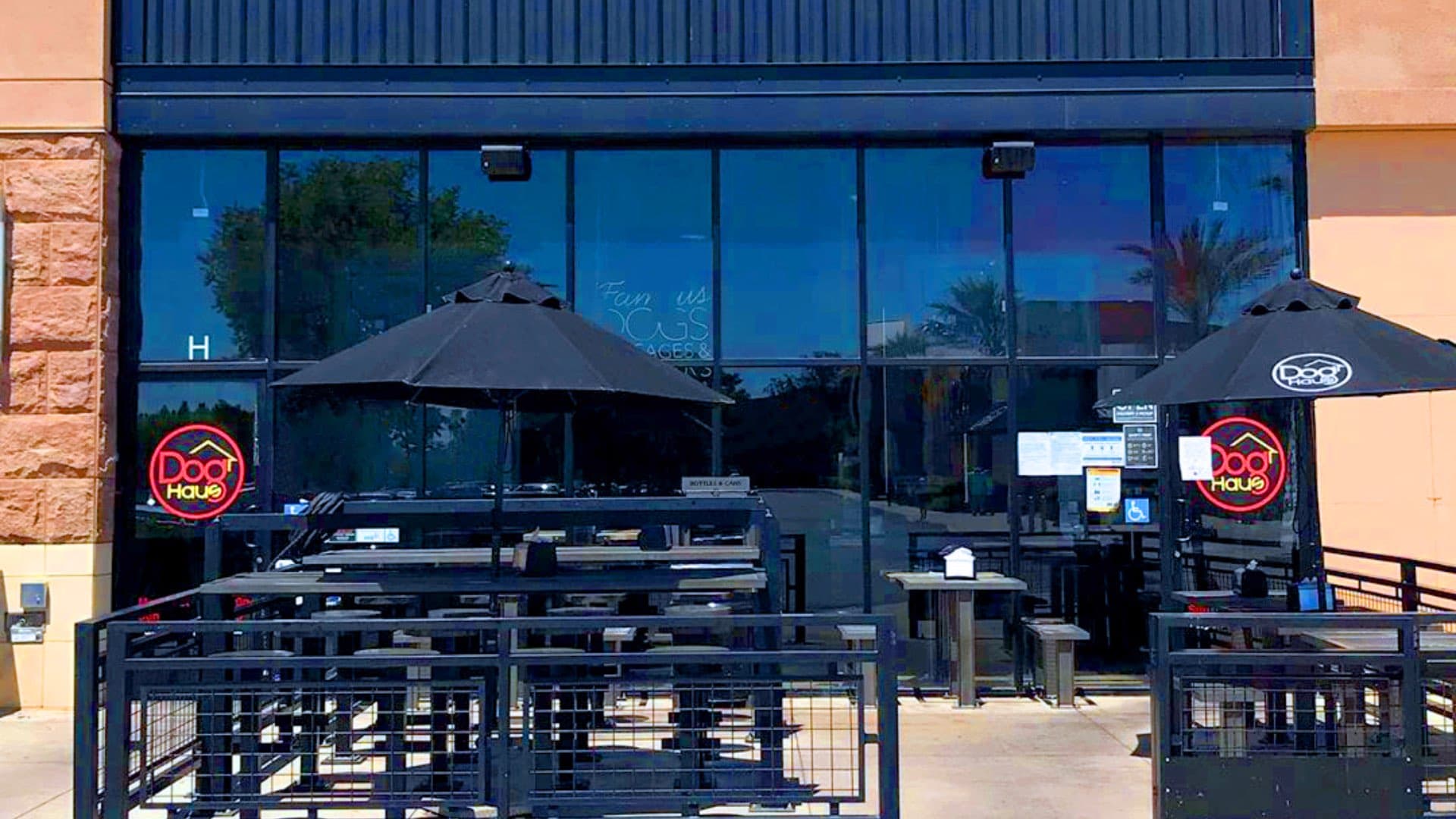 Exterior view of Dog Haus Palmdale with black patio umbrellas and neon logo on glass facade.