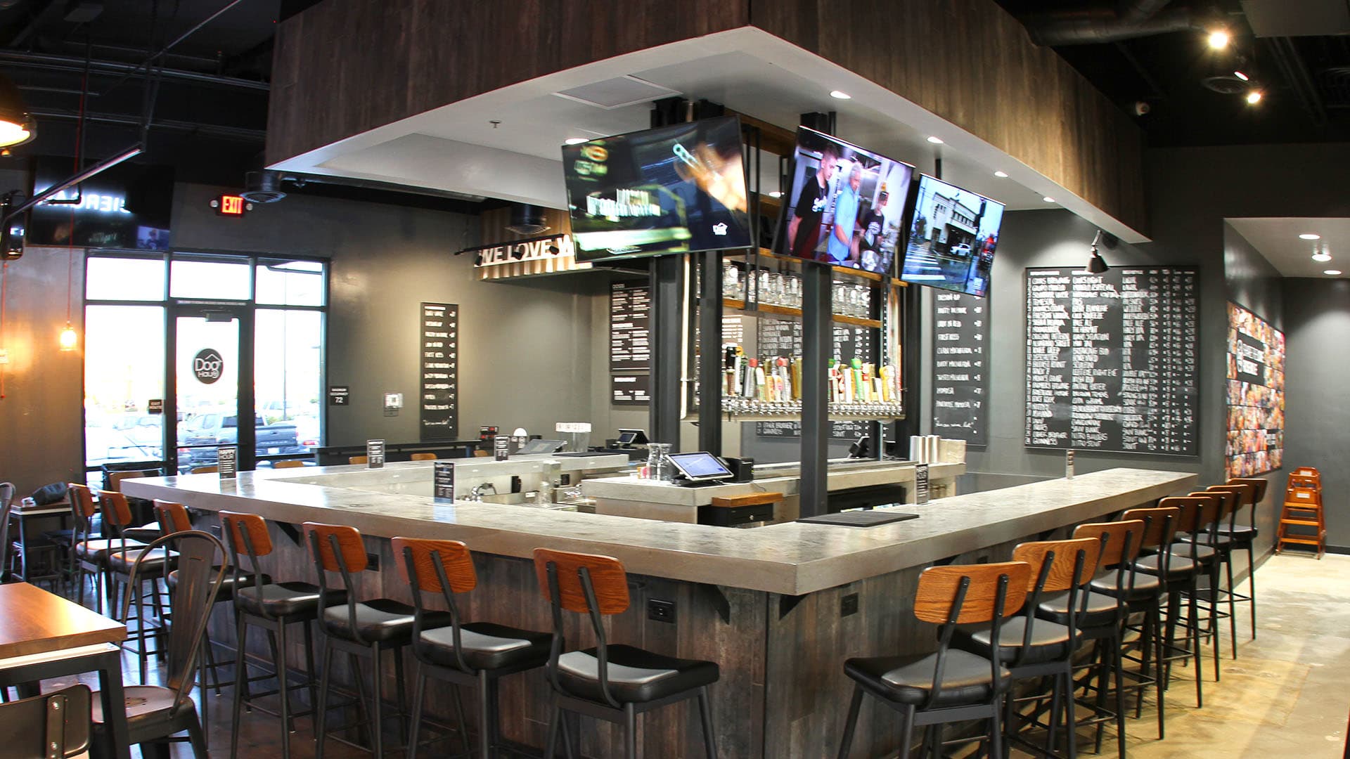 A modern bar with a long counter, multiple bar stools, beer taps, and several TVs mounted above.