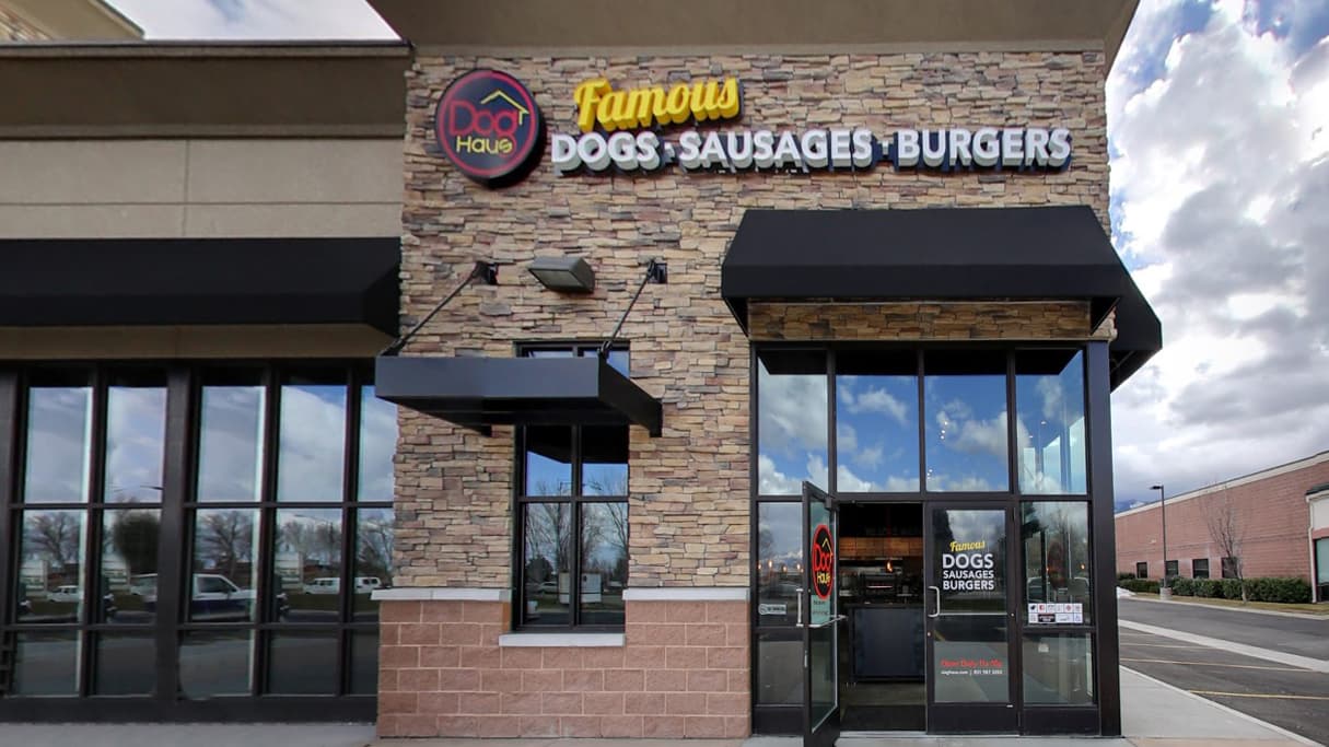 A wide interior view of a Dog Haus restaurant, featuring a large mural on the left wall with "Dog Haus" and "Famous DOGS SAUSAGES BURGERS" text. The dining area has various high-top tables with dark stools and chairs. In the foreground, there's a low black partition. Towards the back right, a counter service area is visible with menu boards above and the text "WE LOVE WIENERS" on the wall. The ceiling has exposed dark pipes and hanging pendant lights.