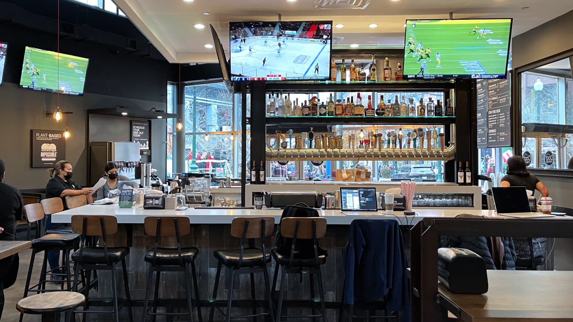 A view of a modern restaurant interior with a bar counter on the right, various tables and stools, and multiple TVs displaying sports.