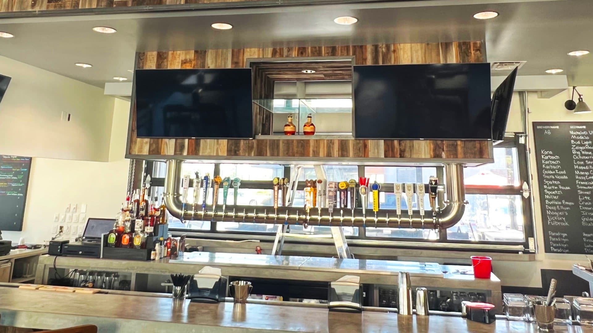 A modern bar with a long row of beer taps, two large TVs, and a wooden paneling backdrop.