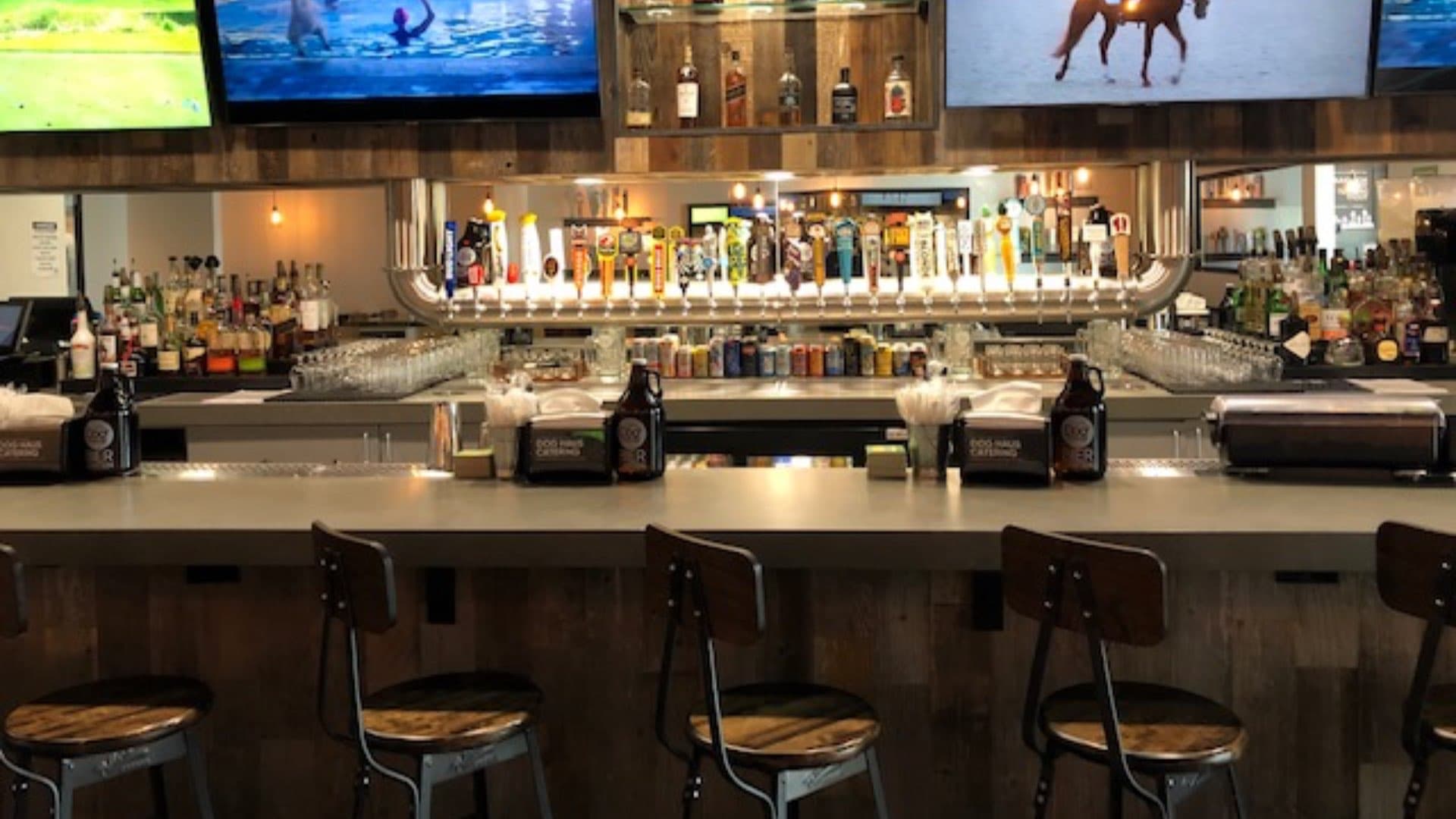 A well-stocked bar with many beer taps, bottles on shelves, and TVs mounted above, with bar stools in the foreground.