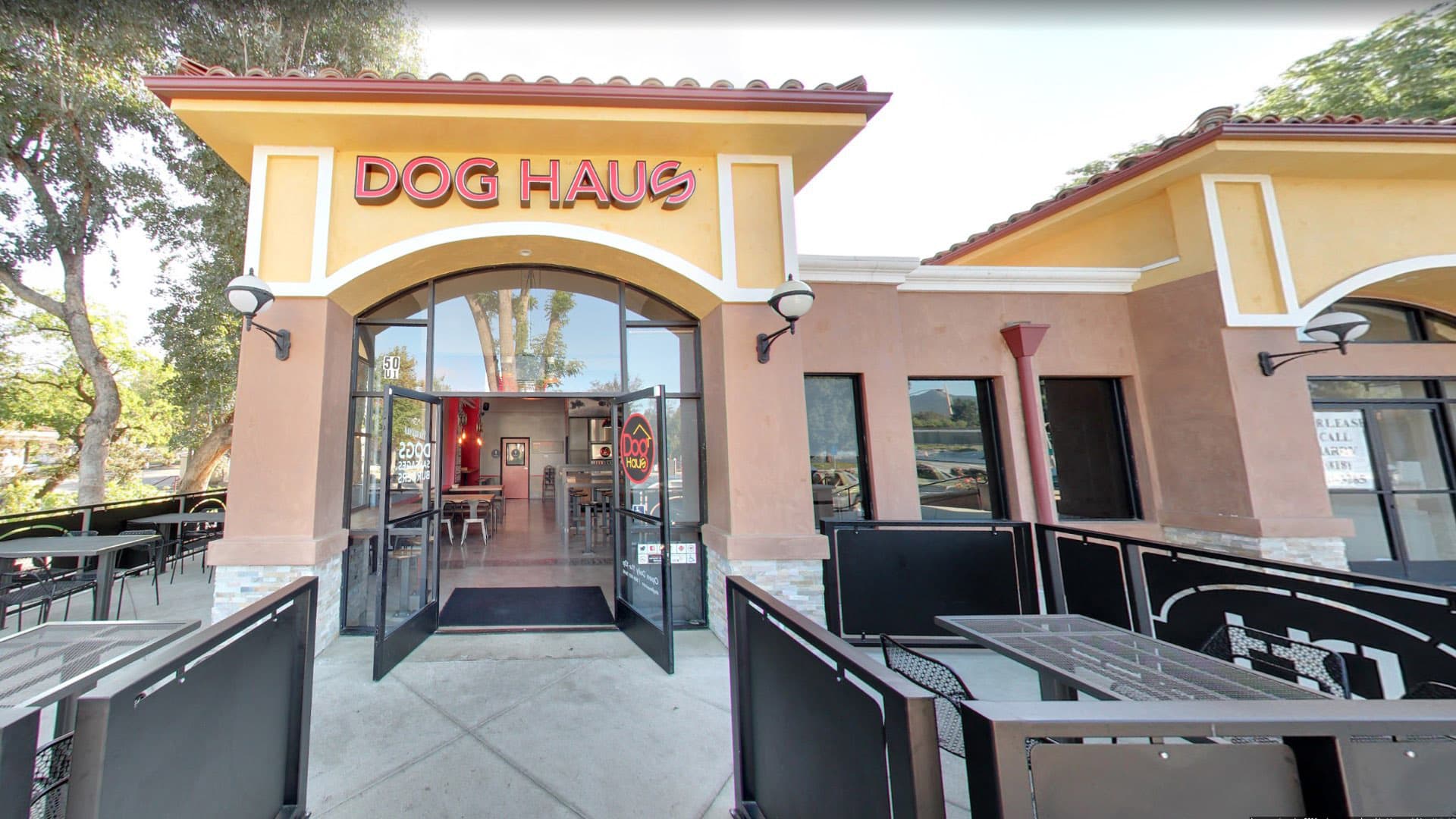The exterior of a "Dog Haus" restaurant with a terracotta roof, a large arched entrance, and outdoor patio seating.