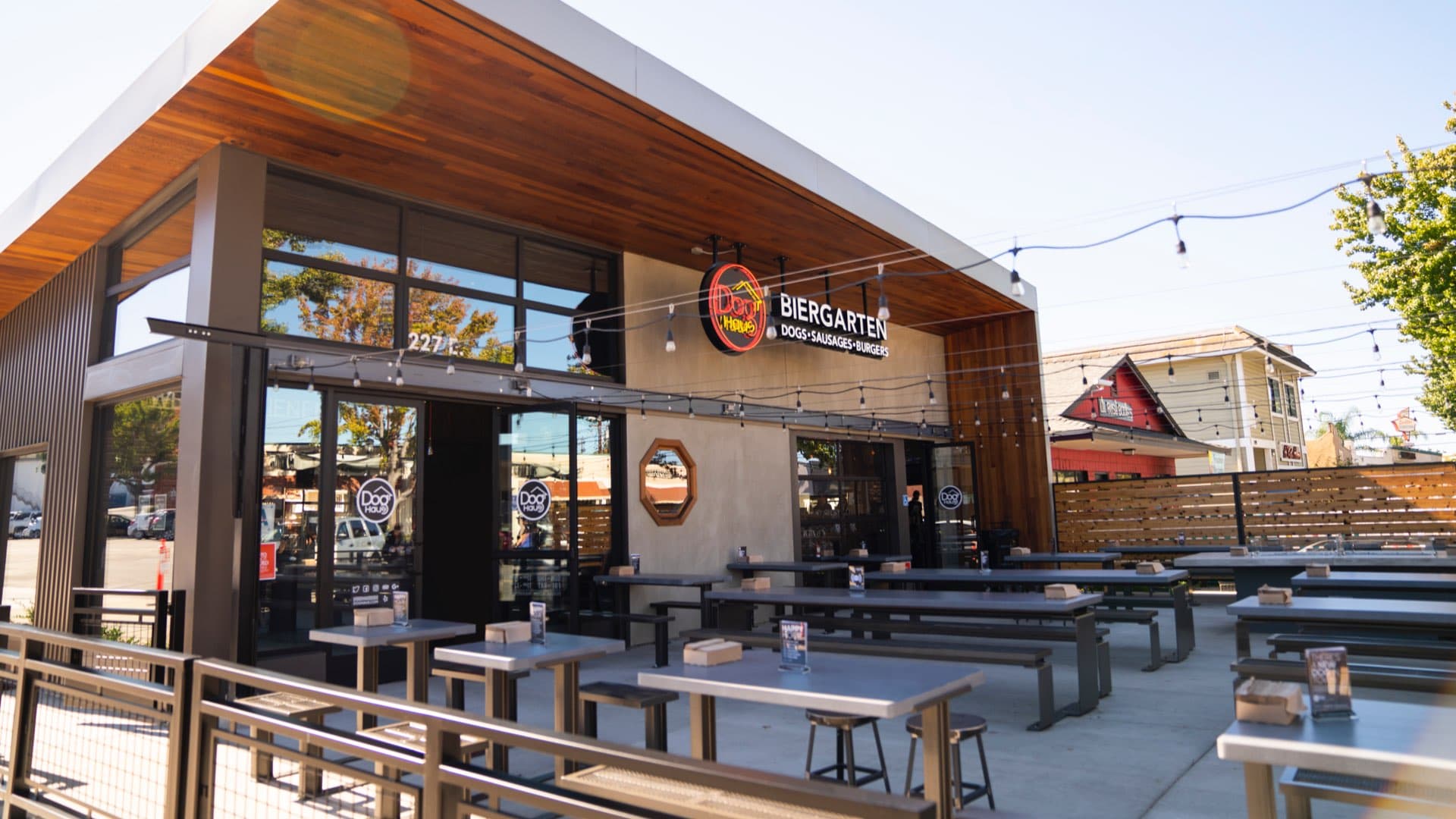 The exterior of a "Dog Haus Biergarten" restaurant with a contemporary design, an expansive outdoor patio with tables, and string lights.