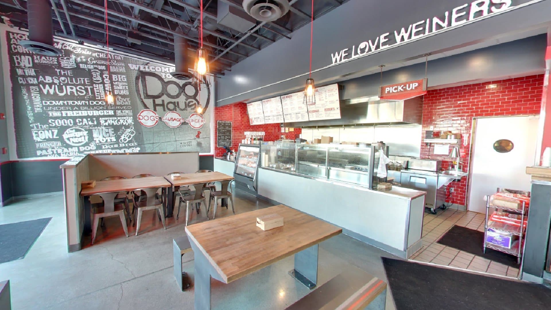 A wide view of a restaurant dining area with booths and tables, leading to a counter service area with "WE LOVE WIENERS" on the wall, and a large chalkboard mural.