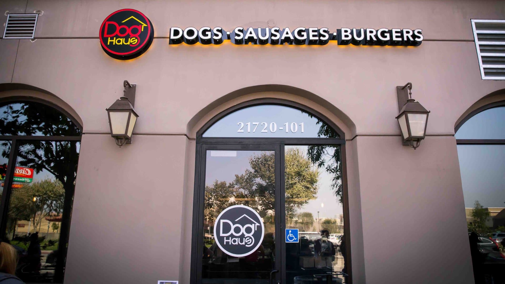 Exterior of Dog Haus Carson featuring arched windows, black door with Dog Haus logo, street number “21720-101” above the entrance, and illuminated signage reading “DOGS • SAUSAGES • BURGERS” mounted above the door.