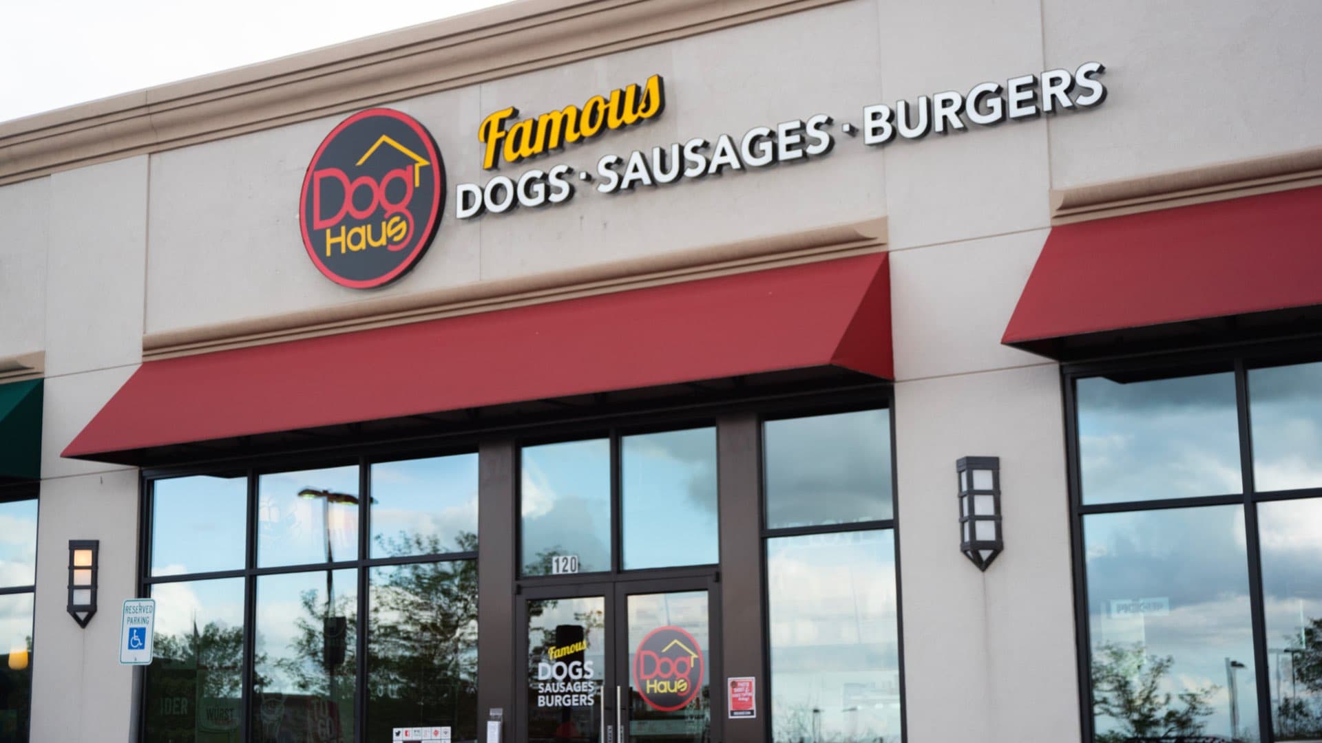 Front view of Dog Haus Centennial with bold signage for hot dogs, sausages, and burgers, red awnings, and a handicap parking sign near the entrance
