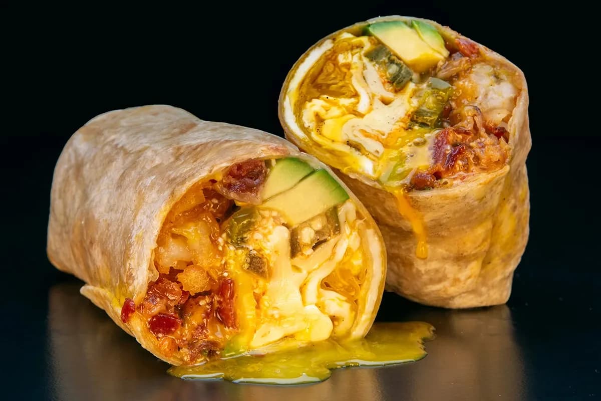 A close-up of two breakfast burrito halves filled with melted cheese, crispy bacon, avocado slices, pickled jalapenos, and a soft-boiled egg with a runny yolk spilling onto a dark surface.