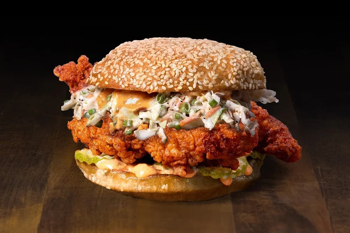 A crispy fried chicken sandwich on a toasted sesame seed bun, topped with creamy coleslaw, sliced pickles, and a spicy signature sauce.