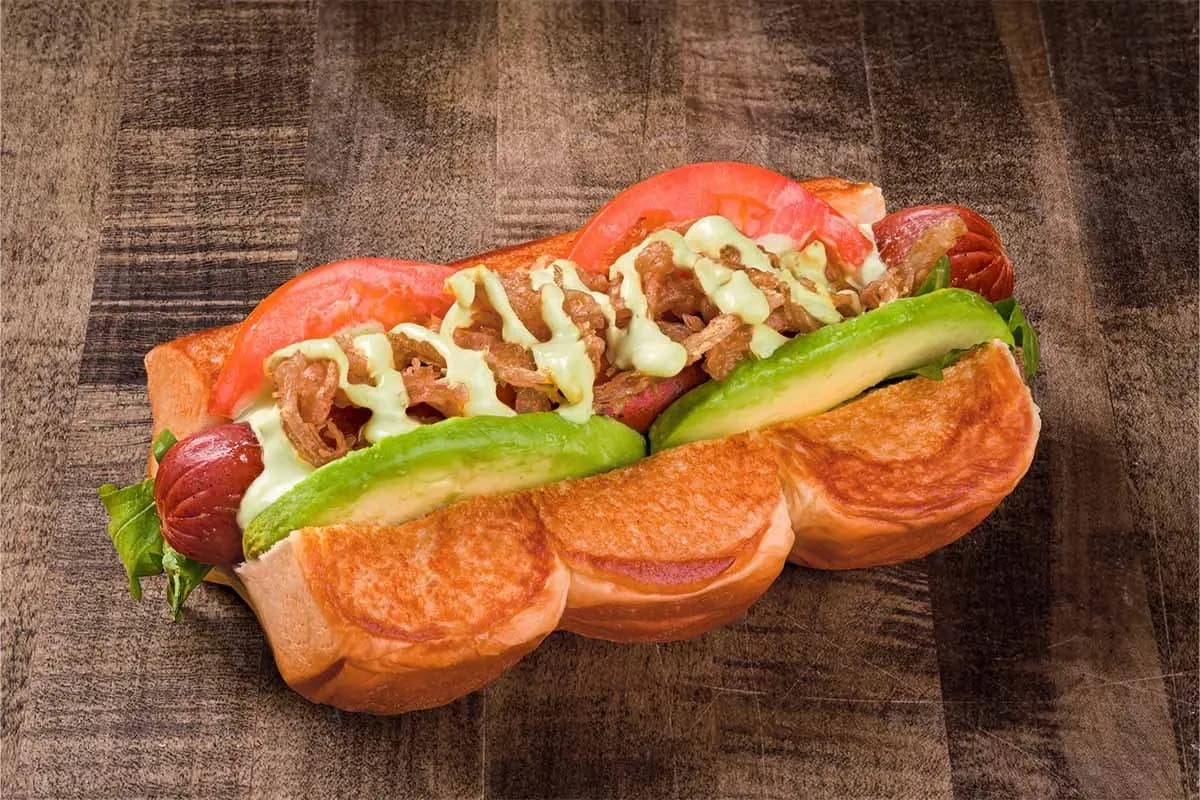 A gourmet hot dog served on a toasted king's Hawaiian-style bun, topped with fresh avocado slices, tomato wedges, crispy onions, and a drizzle of creamy green herb sauce.