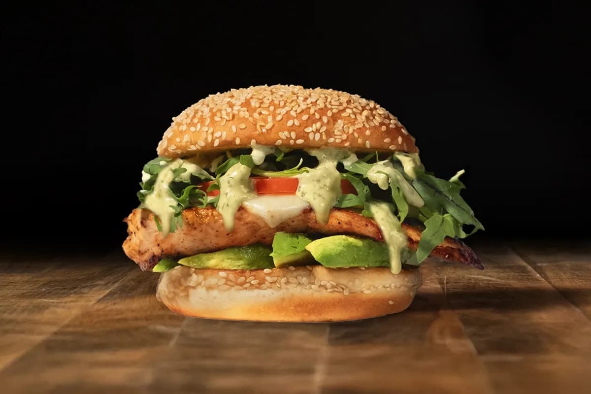 A grilled chicken breast sandwich on a toasted sesame seed bun, layered with fresh avocado, sliced tomato, wild arugula, and a generous drizzle of green herb aioli.
