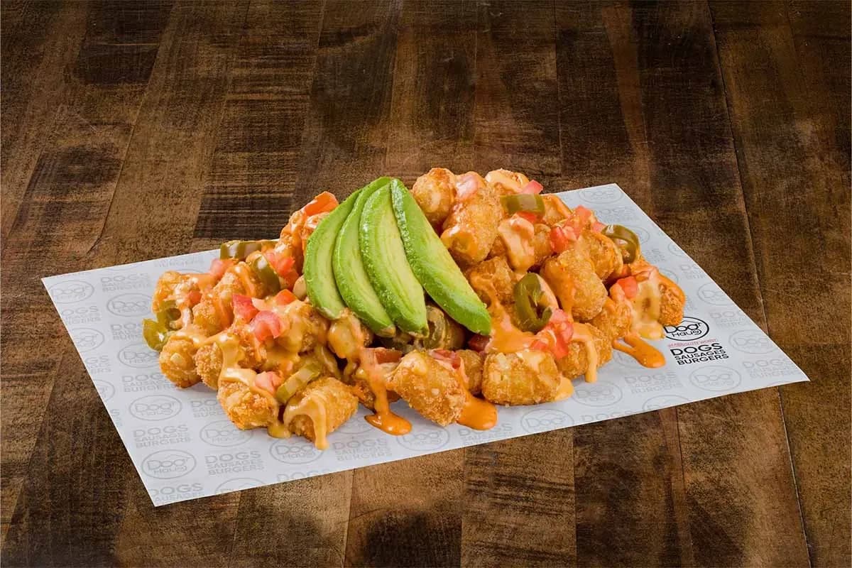 A large portion of crispy tater tots served on branded parchment paper, smothered in melted cheese sauce, diced tomatoes, jalapenos, and topped with several fresh avocado slices.