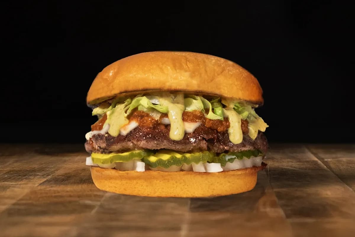 A juicy smashburger with a seared beef patty on a toasted bun, topped with melted cheese, pickles, diced white onions, shredded lettuce, and a signature creamy sauce.