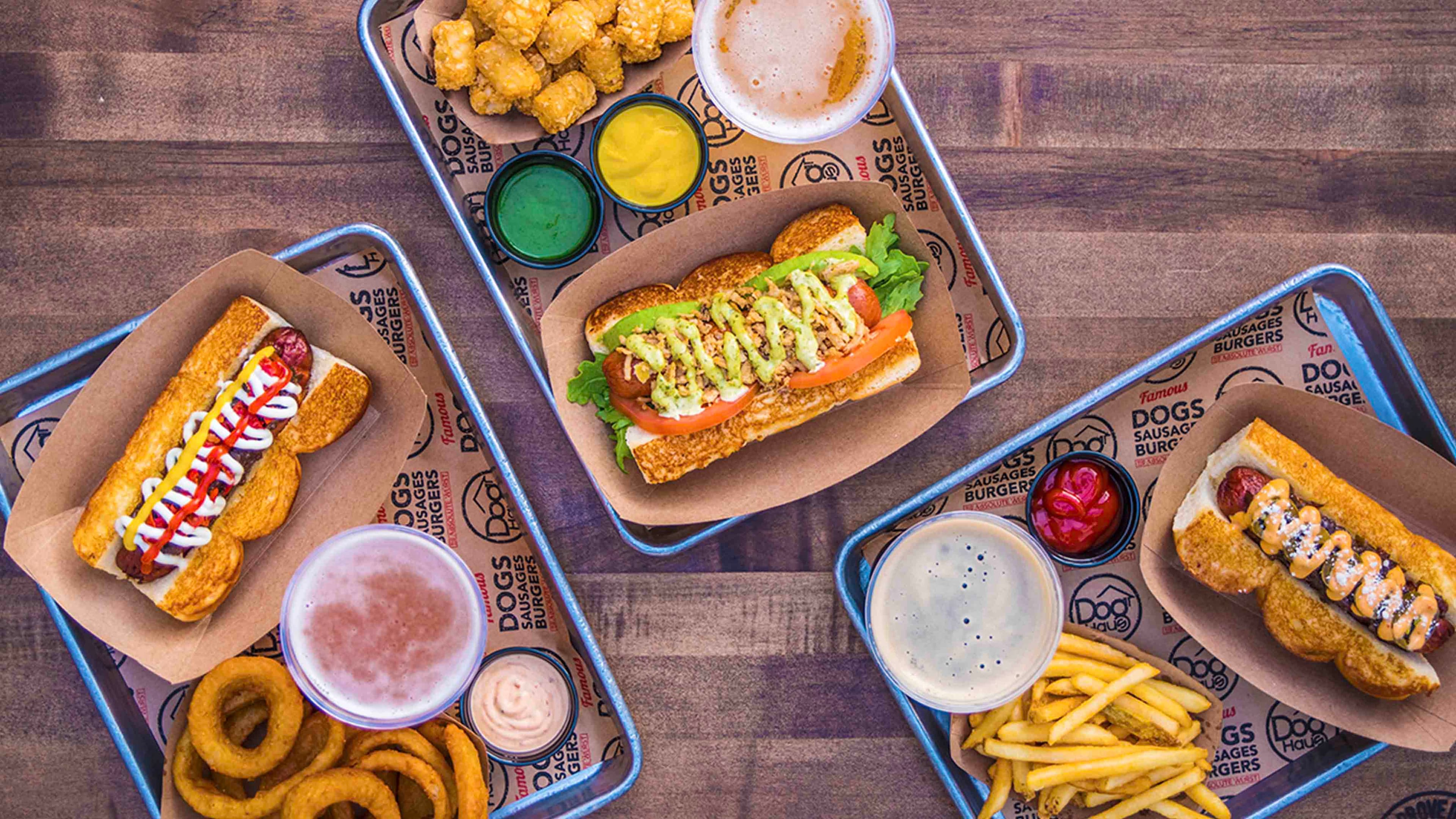 An overhead view of multiple Dog Haus trays filled with gourmet hot dogs, tater tots, onion rings, French fries, and dipping sauces, served on branded paper with beers on a wooden table.