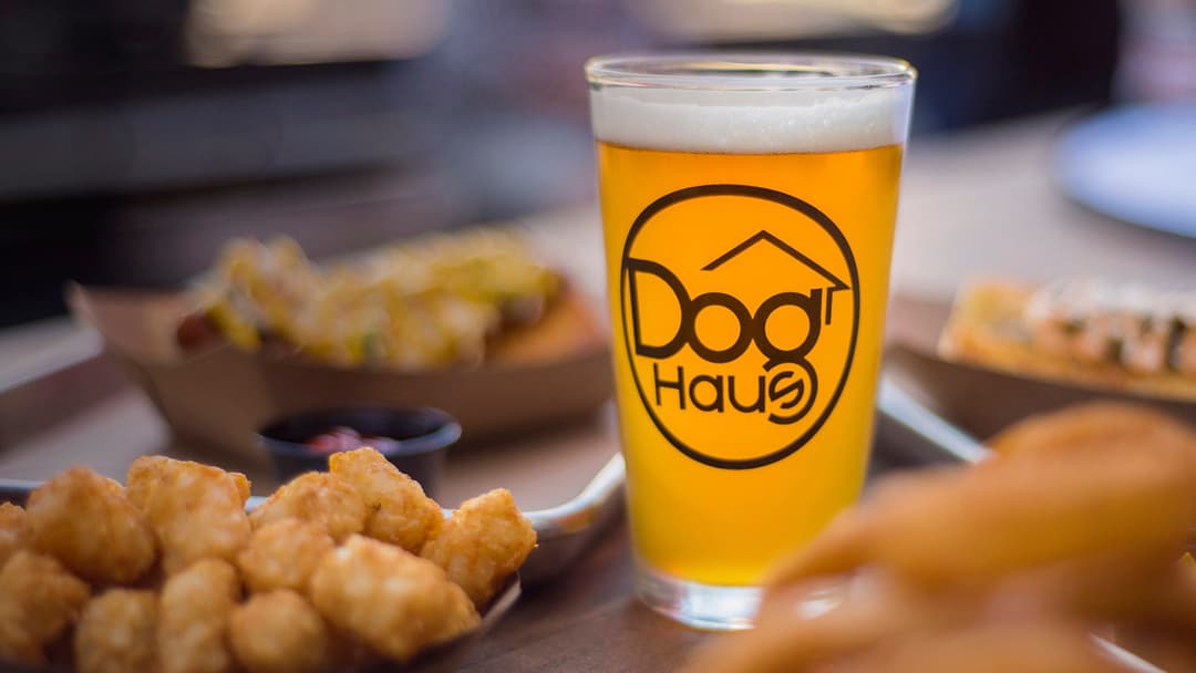 Dog Haus Happy Hour beer with tater tots and hot dogs in the background