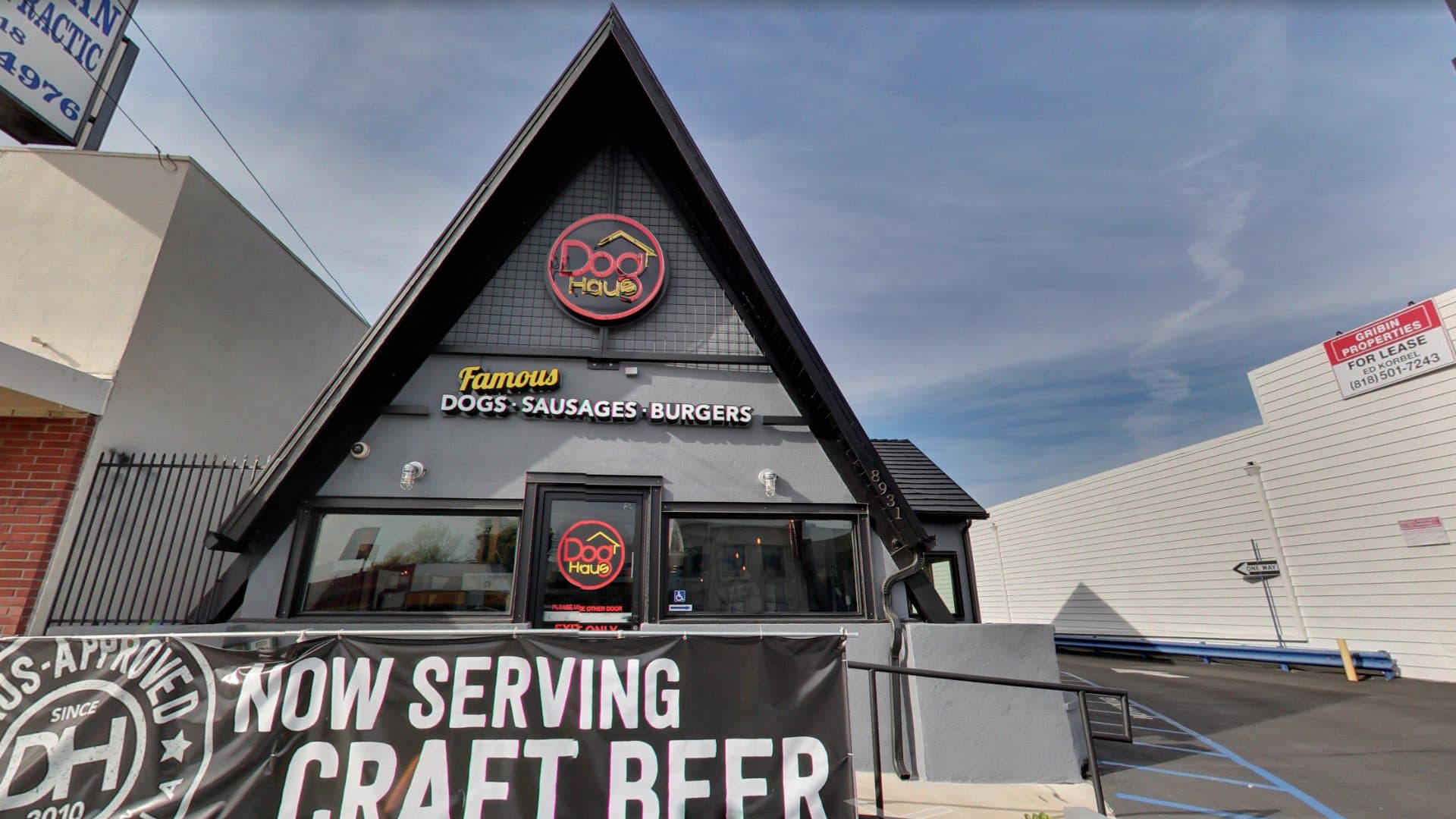 Exterior of Dog Haus Northridge with an A-frame roof, black and gray facade, and signage that reads 'Famous Dogs • Sausages • Burgers' above the entrance.