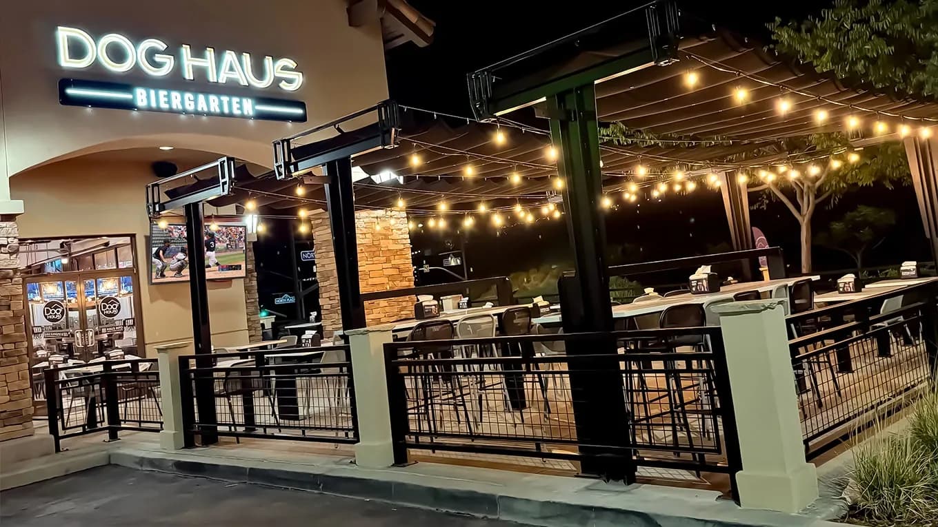 The exterior of Dog Haus Biergarten at night, with an illuminated sign and a covered, string-lit patio filled with tables and chairs.